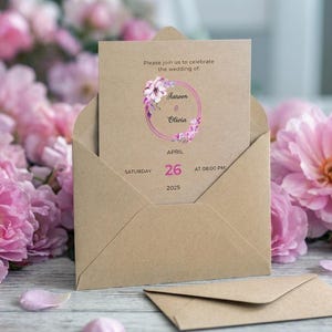 May include: A tan kraft paper envelope containing a matching wedding invitation card. The card features a pink floral wreath graphic, the text Please join us to celebrate the wedding of Aareen & Olivia, and the date Saturday April 26 2025 at 08:00 PM.