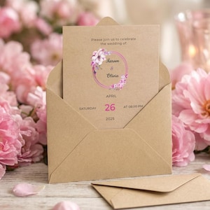 May include: A tan kraft paper wedding invitation card inside a matching envelope. The card features a pink floral wreath graphic, the text 'Please join us to celebrate the wedding of: Aareen & Olivia', and the date 'Saturday April 26 2025'.