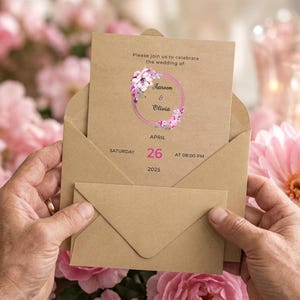 May include: A tan cardstock wedding invitation inside a matching envelope. The text reads: Please join us to celebrate the wedding of: Aaron & Olivia, Saturday, April 26, 2025, at 08:00 PM. A circular floral wreath in shades of pink and purple surrounds the names.
