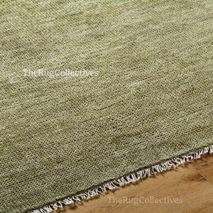 May include: Close-up of a textured, olive green rug with a woven pattern. The rug has a fringe border along one edge and is set on a wooden floor. The Rug Collectives is printed on the rug.