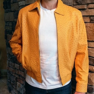 May include: A golden-yellow leather jacket with a textured surface and a zipper closure. The jacket has a collared neckline and is worn over a white t-shirt and blue jeans. The jacket's design includes a ribbed hem and cuffs.