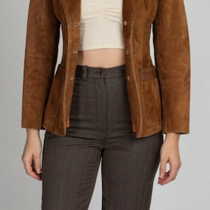May include: A brown suede blazer with a notched lapel and two front pockets. The blazer is paired with brown trousers. The outfit is completed with a cream-colored top. The blazer has a classic, tailored fit.