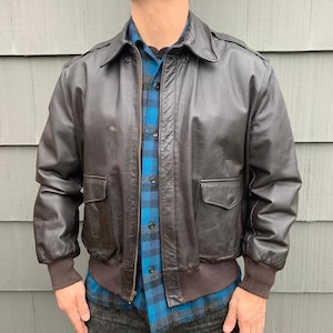 Puede incluir: Chaqueta bomber de cuero marrón oscuro con cremallera, dos bolsillos delanteros y dobladillo y puños de canalé. La chaqueta tiene un cuello clásico y charreteras en los hombros. La chaqueta se lleva sobre una camisa a cuadros azul y negro.