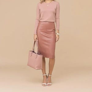 May include: A woman's outfit in a monochromatic color scheme. The outfit includes a long-sleeved, V-neck sweater, a knee-length pencil skirt, a tote bag, and high-heeled sandals. The color palette is a soft, muted pink.