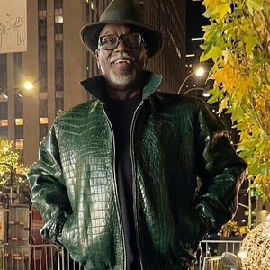 May include: A man wearing a dark green, textured leather jacket with a black shirt and a dark green hat. The jacket has a zipper closure and a collar. The background includes a city street with buildings and trees.