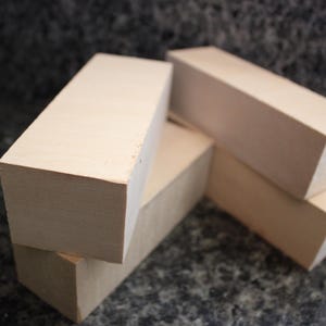 May include: A collection of unfinished, rectangular wooden blocks. The blocks are stacked and arranged, showcasing their natural, light-colored wood grain. The edges are clean and sharp, suggesting they are freshly cut.