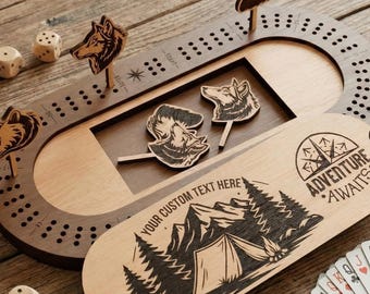 Peut inclure: Un jeu de cribbage en bois avec un couvercle décoratif représentant une montagne et une tente, les mots "Adventure Awaits" et le texte "Your Custom Text Here". Le jeu comprend des chevilles en forme d'animaux, des dés et un jeu de cartes.