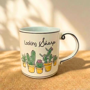 Puede incluir: Una taza de cerámica blanca con borde y asa negros. La taza presenta una ilustración dibujada a mano de cactus en macetas amarillas, con las palabras "Looking Sharp" en escritura cursiva. La taza está sobre un fondo neutro.