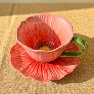 May include: A ceramic teacup and saucer set designed to resemble a poppy flower. The cup and saucer are shades of pink and red, with a green handle. The cup's interior has a yellow center.