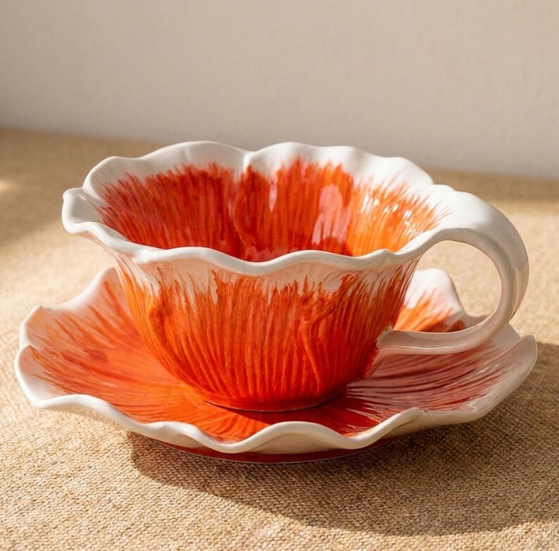 Puede incluir: Un juego de taza y platillo de cer&aacute;mica. La taza y el platillo tienen forma de flor con borde y asa blancos. El interior de la taza y el platillo es naranja con pinceladas blancas. El juego est&aacute; sobre una superficie de color neutro.