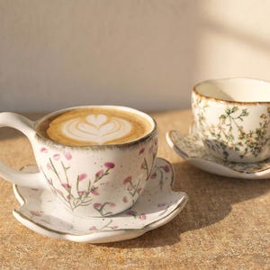 Puede incluir: Dos tazas de café de cerámica con platillos. Una taza está llena de arte latte. Ambas tazas y platillos son blancos con un estampado floral en rosa y verde. Los platillos tienen forma de hoja.