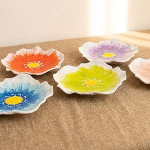 Puede incluir: Un conjunto de seis platos de cer&aacute;mica con forma de flor. Cada plato presenta una combinaci&oacute;n de colores diferente, incluyendo naranja, azul, verde, morado y rosa, con un centro amarillo. Los platos est&aacute;n dispuestos sobre una superficie de tono neutro.
