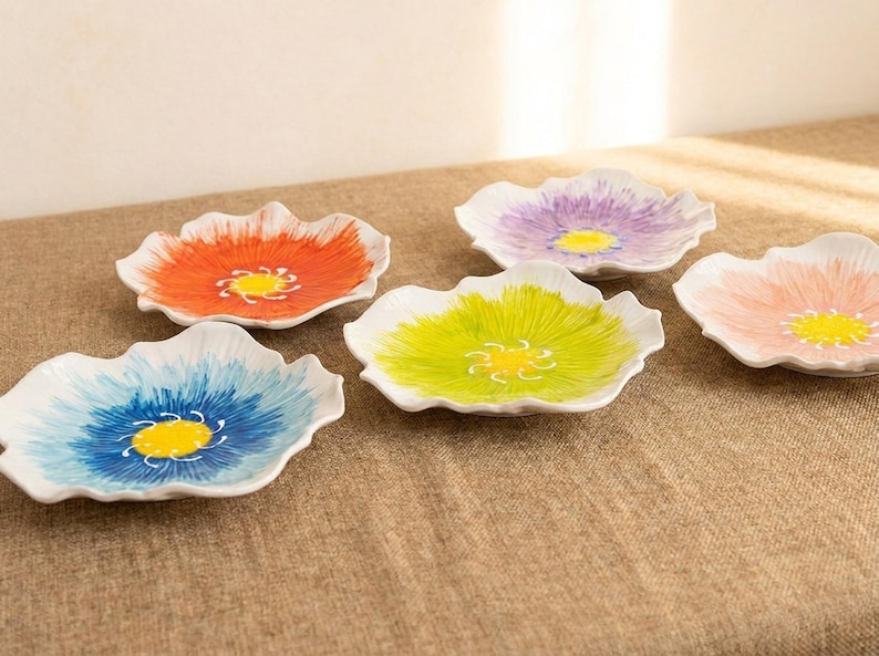 Puede incluir: Un conjunto de seis platos de cer&aacute;mica con forma de flor. Cada plato presenta una combinaci&oacute;n de colores diferente, incluyendo naranja, azul, verde, morado y rosa, con un centro amarillo. Los platos est&aacute;n dispuestos sobre una superficie de tono neutro.