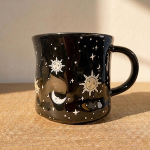 May include: Black ceramic mug with a celestial design. The mug features white illustrations of the sun, moon, and stars. The mug has a curved shape and a black handle.