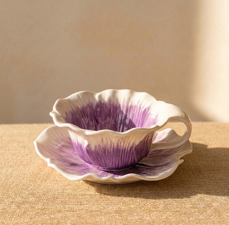 Puede incluir: Un juego de taza y platillo de cer&aacute;mica. La taza y el platillo tienen forma de flor con p&eacute;talos blancos y morados. El juego est&aacute; sobre una superficie beige.