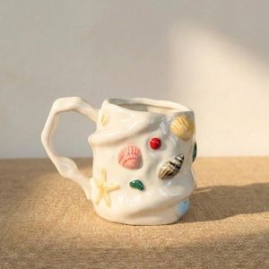 以下が含まれることがあります： クリーム色の斑点のある釉薬が施された、3つのセラミックマグのセット。各マグには、青と緑の葉、黄色の花、赤いベリーなど、異なる手描きのデザインが施されています。マグはシンプルでモダンなデザインです。