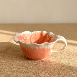 Puede incluir: Una taza de t&eacute; de cer&aacute;mica con un dise&ntilde;o floral. La taza es principalmente de color coral con detalles blancos en el borde y el asa. La forma de la taza se asemeja a una flor, con un borde festoneado.