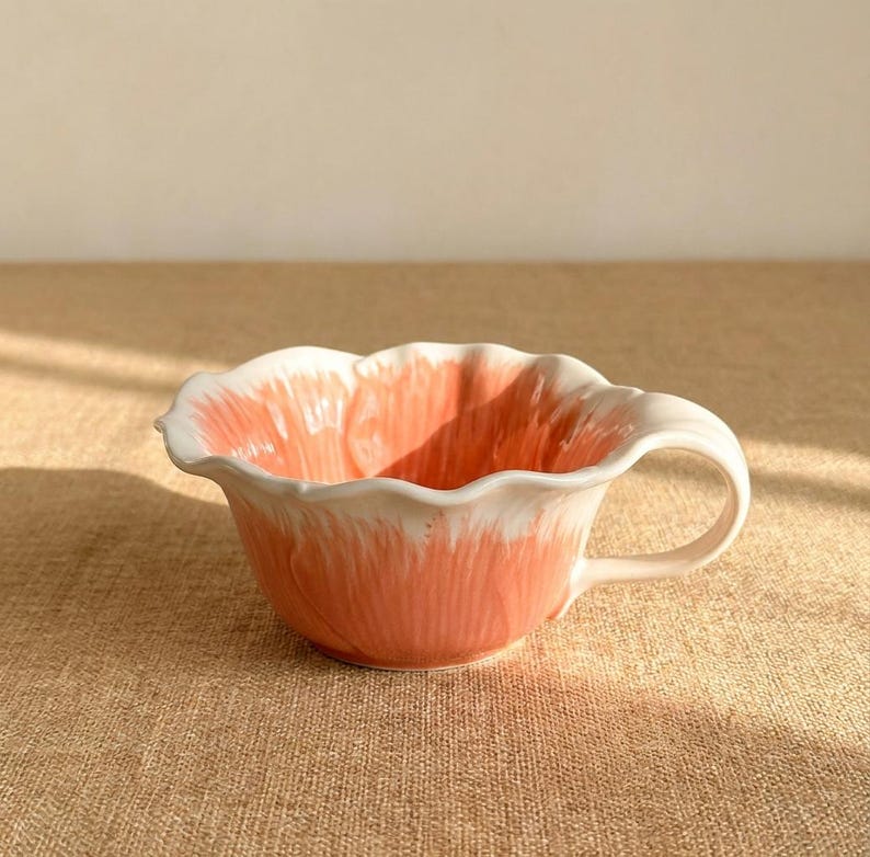 Puede incluir: Una taza de t&eacute; de cer&aacute;mica con un dise&ntilde;o floral. La taza es principalmente de color coral con detalles blancos en el borde y el asa. La forma de la taza se asemeja a una flor, con un borde festoneado.