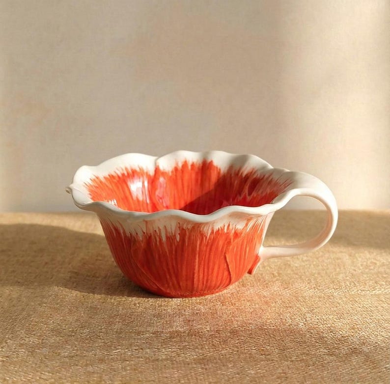 Puede incluir: Una taza de t&eacute; de cer&aacute;mica con un dise&ntilde;o floral. La taza es principalmente naranja con detalles blancos en el borde y el asa. La taza est&aacute; sobre una superficie beige.
