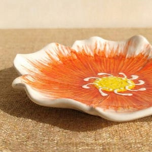 Puede incluir: Un plato de cer&aacute;mica con forma de flor, con borde blanco y centro naranja vibrante. El centro presenta un dise&ntilde;o floral amarillo y blanco. El plato est&aacute; sobre una superficie texturizada de color beige.