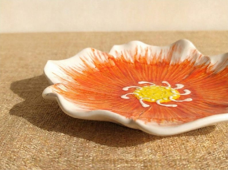 Puede incluir: Un plato de cer&aacute;mica con forma de flor, con borde blanco y centro naranja vibrante. El centro presenta un dise&ntilde;o floral amarillo y blanco. El plato est&aacute; sobre una superficie texturizada de color beige.