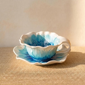 Puede incluir: Un juego de taza y platillo de cer&aacute;mica. La taza y el platillo tienen forma de flor con un dise&ntilde;o de acuarela blanco y azul. La taza tiene un asa y se apoya en el platillo a juego. El conjunto est&aacute; sobre una superficie de color neutro.