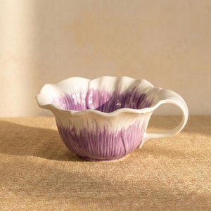 Puede incluir: Una taza de t&eacute; de cer&aacute;mica con un dise&ntilde;o floral. La taza tiene forma de flor, con un borde blanco y p&eacute;talos morados. Tiene un asa curva y est&aacute; sobre un fondo neutro.