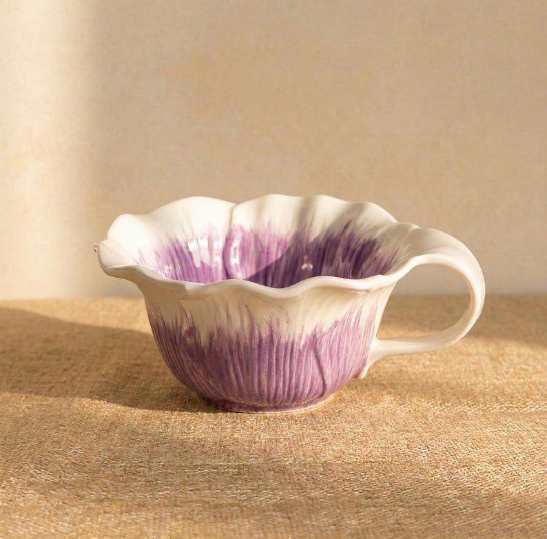 Puede incluir: Una taza de t&eacute; de cer&aacute;mica con un dise&ntilde;o floral. La taza tiene forma de flor, con un borde blanco y p&eacute;talos morados. Tiene un asa curva y est&aacute; sobre un fondo neutro.