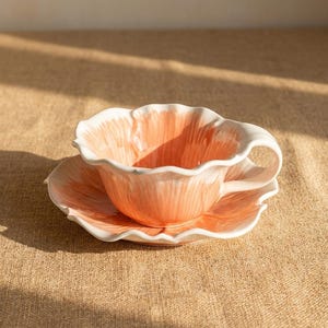 Puede incluir: Un juego de taza y platillo de cer&aacute;mica. La taza y el platillo tienen forma de flor con interior color melocot&oacute;n y ribete blanco. El juego est&aacute; sobre una superficie de color neutro.