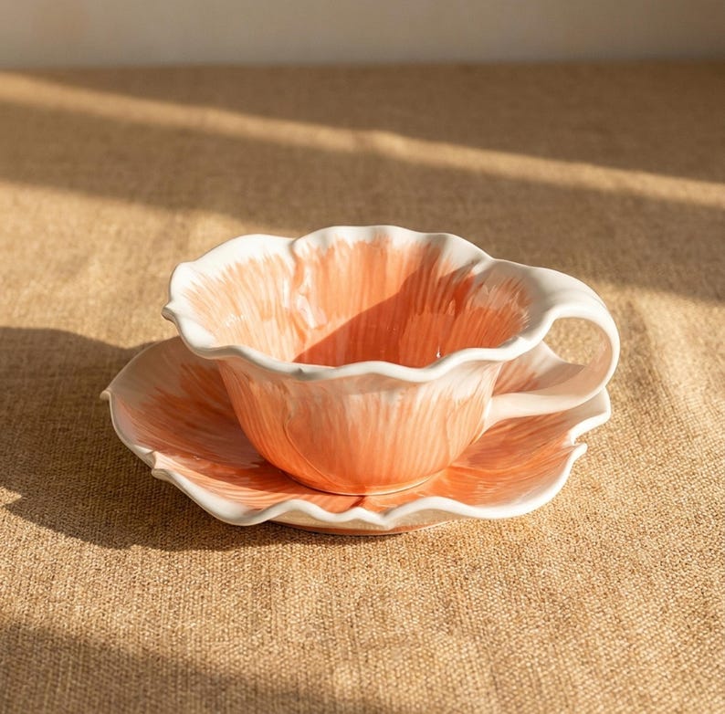 Puede incluir: Un juego de taza y platillo de cer&aacute;mica. La taza y el platillo tienen forma de flor con interior color melocot&oacute;n y ribete blanco. El juego est&aacute; sobre una superficie de color neutro.