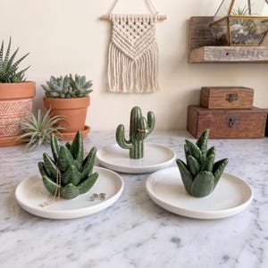 May include: Three white ceramic ring dishes, each featuring a green glazed succulent or cactus figurine in the center. One dish holds a gold chain necklace and small stud earrings, another holds a ring on the cactus, and the third is empty.