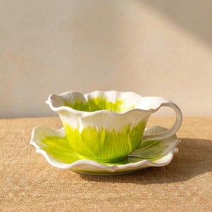 Puede incluir: Un juego de taza y platillo de cer&aacute;mica blanca y verde. La taza y el platillo tienen forma de flor, con exterior blanco e interior verde. El juego est&aacute; sobre una superficie de color neutro.