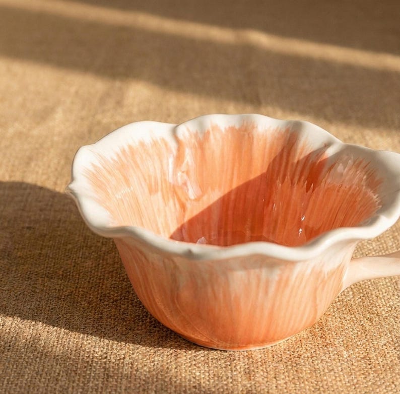 Puede incluir: Una taza de t&eacute; de cer&aacute;mica con un dise&ntilde;o floral. La taza presenta un interior y exterior de color melocot&oacute;n con detalles blancos en el borde. El asa es de color melocot&oacute;n claro. La taza est&aacute; sobre una superficie marr&oacute;n claro.