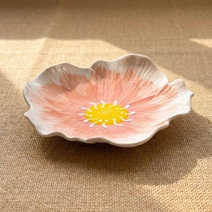 Puede incluir: Un plato de cer&aacute;mica con forma de flor con un dise&ntilde;o pintado en rosa y blanco. El centro del plato es amarillo con detalles blancos. El plato est&aacute; sobre una superficie de color neutro.