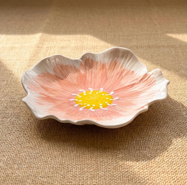 Puede incluir: Un plato de cer&aacute;mica con forma de flor con un dise&ntilde;o pintado en rosa y blanco. El centro del plato es amarillo con detalles blancos. El plato est&aacute; sobre una superficie de color neutro.