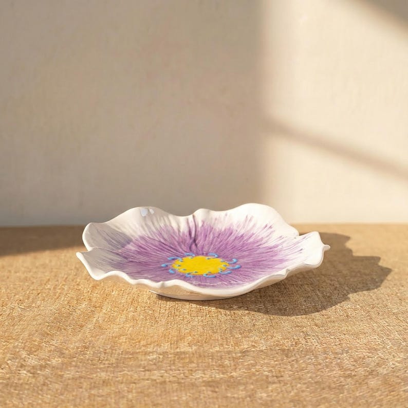 Puede incluir: Un plato de cer&aacute;mica blanco con forma de flor, con un centro morado y detalles amarillos. El plato tiene un borde ondulado y est&aacute; sobre una superficie de madera. El dise&ntilde;o floral es detallado y decorativo.