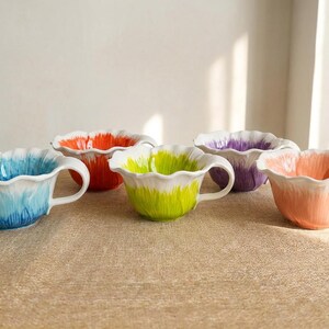 Puede incluir: Conjunto de cinco tazas de t&eacute; de cer&aacute;mica con dise&ntilde;o floral. Cada taza presenta un degradado de color &uacute;nico: azul, naranja, verde, morado y melocot&oacute;n, con detalles blancos. Las tazas tienen asa y borde festoneado.