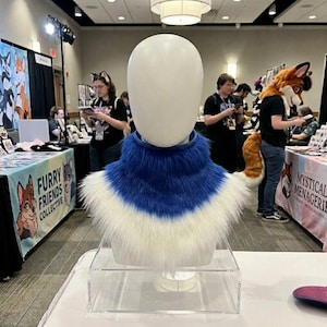 May include: A blue, white, and blue faux fur neck piece displayed on a mannequin head. The furry accessory is likely for a costume or cosplay. The background shows a convention setting with tables and people.