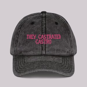 May include: A distressed black baseball cap with a curved brim. The cap features the embroidered text "THEY CASTRATED CASTRO" in pink, centered on the front. The hat has a vintage, worn look.