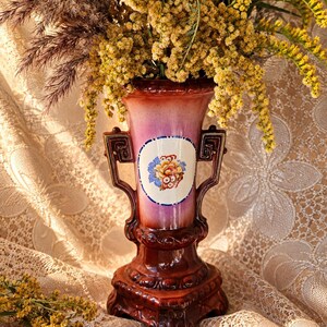 Puede incluir: Un jarrón de cerámica con acabado degradado en rosa y marrón, con dos asas ornamentadas y un medallón central blanco con diseño floral. El jarrón está lleno de flores de vara de oro amarillas y hierbas secas marrones sobre una base cuadrada.