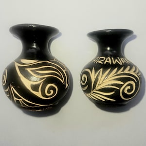 Puede incluir: Dos pequeños jarrones de cerámica negra con cuerpo bulboso y borde acampanado. Cada jarrón presenta un diseño tallado de color crema con hojas arremolinadas y la palabra "ARAWAK" en uno. Cerámica decorativa.