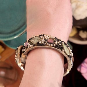 May include: A gold-coloured hinged bracelet with a decorative band. The band is adorned with small black and white beads, along with green, pink, and brown stones. The bracelet is worn on a person's wrist.