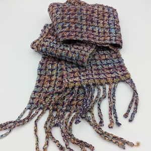 May include: A handwoven scarf in a textured weave, featuring a mix of purple, blue, and gold threads. The scarf has a fringed edge, adding a decorative element. This accessory is designed to be worn around the neck.