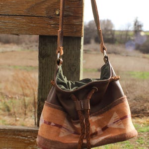 Puede incluir: Bolso de hombro de cuero marr&oacute;n y tela estampada con cierre de cord&oacute;n trenzado. El bolso presenta una correa de ante y un estampado decorativo en tonos naranja y marr&oacute;n. El bolso cuelga de un poste de valla de madera.