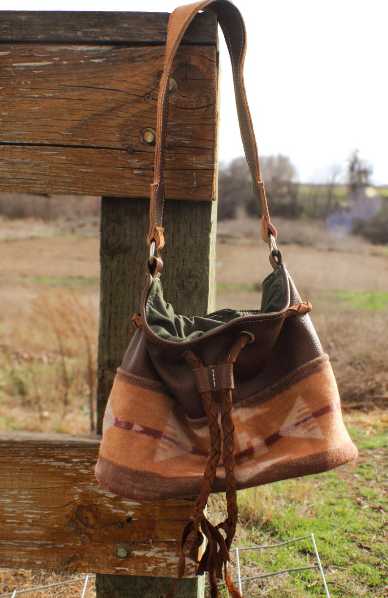 Puede incluir: Bolso de hombro de cuero marr&oacute;n y tela estampada con cierre de cord&oacute;n trenzado. El bolso presenta una correa de ante y un estampado decorativo en tonos naranja y marr&oacute;n. El bolso cuelga de un poste de valla de madera.