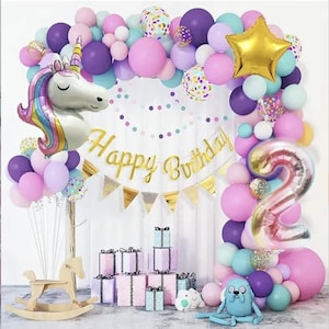 Puede incluir: Una escena festiva de cumpleaños con un globo de unicornio, un globo de estrella dorada y un globo con el número 2. La escena incluye una pancarta de "Happy Birthday", globos coloridos en tonos rosa, morado y turquesa, y regalos envueltos. También hay un caballito balancín de madera.