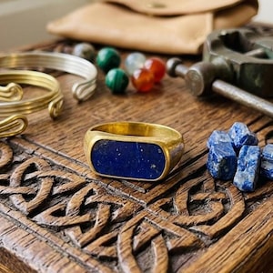 May include: A gold-toned ring featuring a rectangular, deep blue lapis lazuli inlay. The ring is positioned on a dark, carved wooden surface alongside several loose blue lapis lazuli stones, two metal wire bracelets, and a small metal tool.