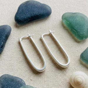 May include: A pair of elongated, oval-shaped silver hoop earrings with a smooth, polished finish. The earrings feature a simple, continuous wire design with a hinged post closure, presenting a clean and minimalist aesthetic for everyday wear.