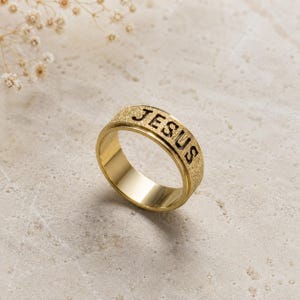 Könnte beinhalten: Ein goldfarbener Ring mit einer texturierten Außenfläche, in die das Wort JESUS in fetten, dunklen Großbuchstaben eingraviert ist. Das Band hat eine glatte, polierte Innenfläche und eine durchgehend gleichmäßige Breite.
