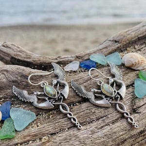May include: A pair of silver-toned earrings featuring a bird with outstretched wings perched above a coiled snake design. Each earring includes a central oval-shaped iridescent gemstone and a curved ear wire for attachment to the earlobe.